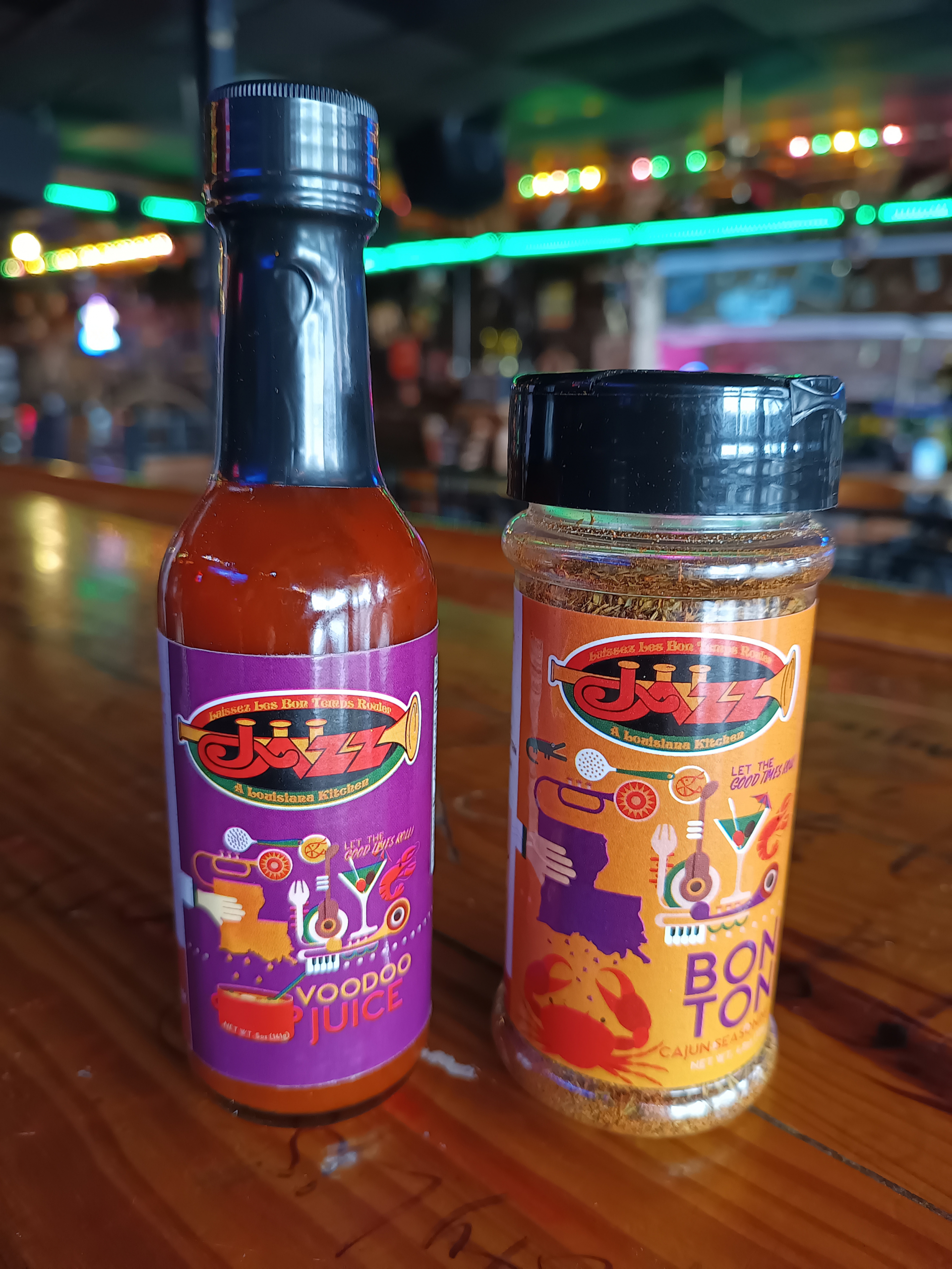 Jazz - A Louisiana Kitchen Two containers on a wooden table: a bottle labeled "Voodoo Juice" and a jar labeled "Bon Ton" with colorful graphics. Background features a bar with lights and decor. Kansas City, Columbia, cajun food restaurant near me