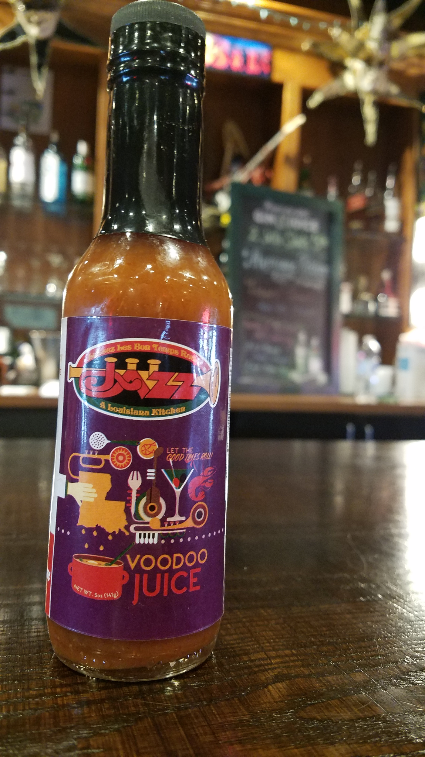 Jazz - A Louisiana Kitchen A bottle of Voodoo Juice hot sauce labeled "Jazz: A Louisiana Kitchen" sits on a wooden surface, with a bar in the background. Kansas City, Columbia, cajun food restaurant near me